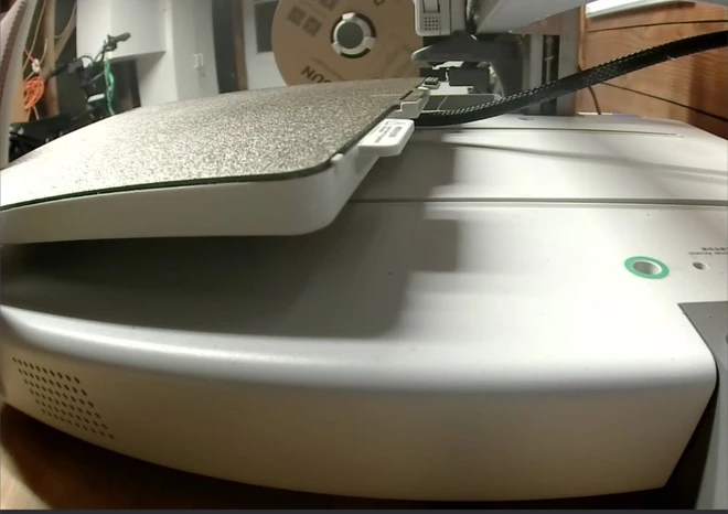A low-angle frame from a 3D printer's own camera showing an empty textured build plate, with a filament spool in the background.