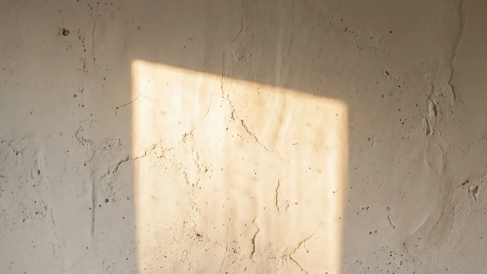 A slanted rectangle of warm sunlight falling across a cracked, pitted plaster wall.