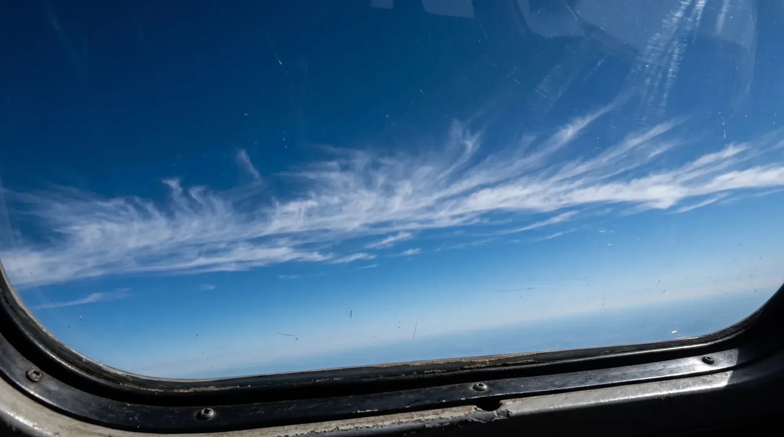 High cirrus clouds over a blue horizon, seen through the curved window of an aircraft cabin.