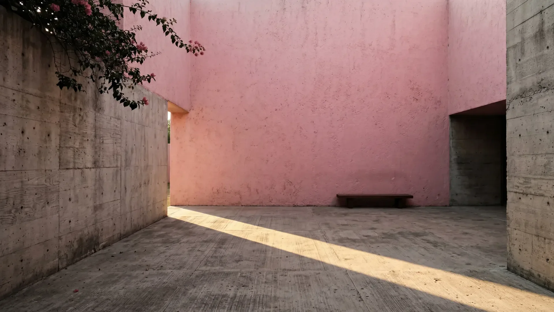 A concrete courtyard enclosed by a tall pink wall, a wooden bench at its foot and a band of late sunlight crossing the floor from an opening in the wall.